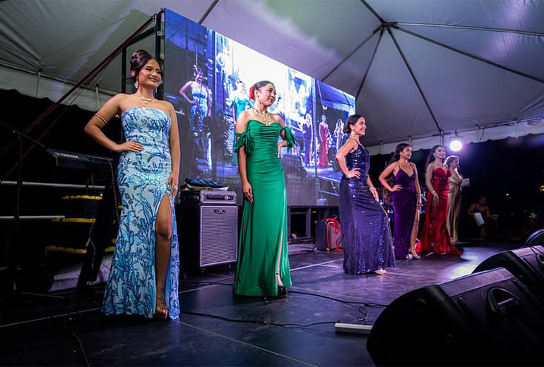 Miss UOG contestants stand in a line on stage in various colored evening gowns.